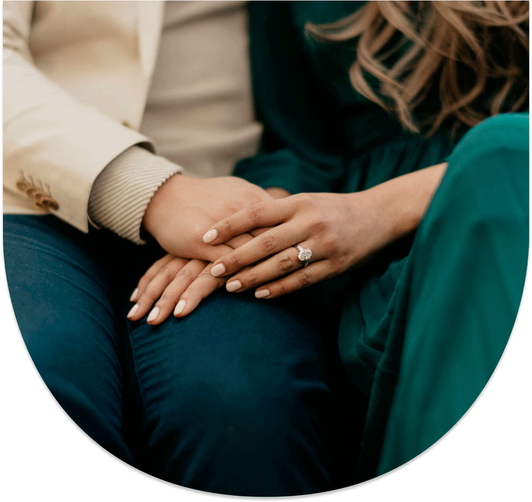 A close-up of two hands resting together, one adorned with a ring, against a backdrop of elegant clothing.